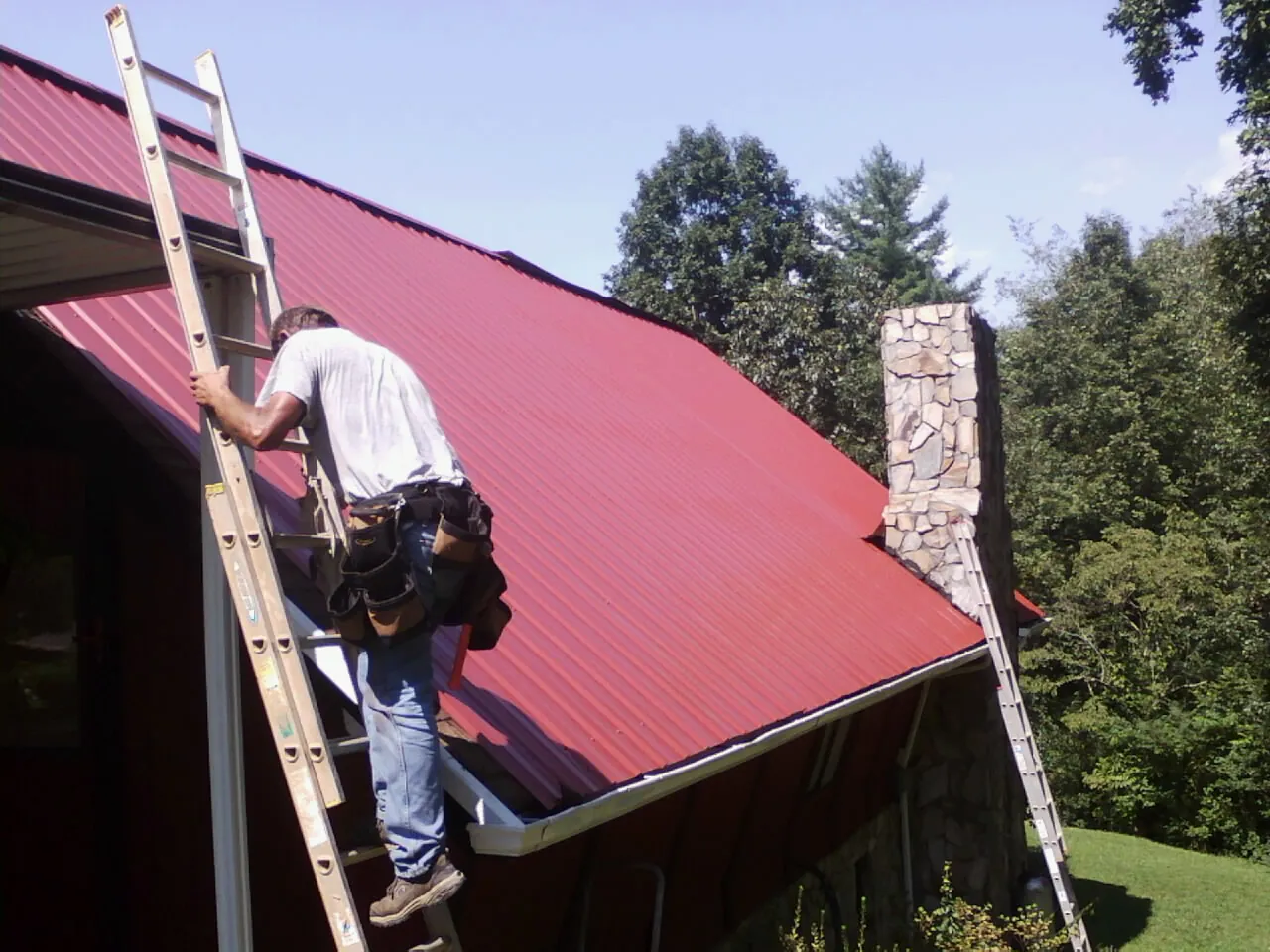 Roofer on a ladder beside a red metal roof and stone chimney — Soffit & Fascia Repair by Roofing Brothers in Point Reyes Station